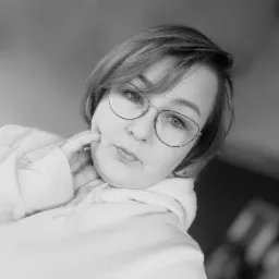 Portret kobiety w okularach, z krótkimi włosami, ubrana w bluzę z kapturem, pozująca z dłonią przy twarzy w czarno-białej tonacji.