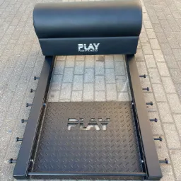 Czarna konstrukcja metalowa z wyściełanym wałkiem i platformą z blachy ryflowanej, oznaczona logo 'PLAY MADE BY ATHLETES', ustawiona na szarej kostce brukowej.