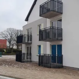 Nowoczesny budynek wielorodzinny z balkonami zabezpieczonymi czarnymi, metalowymi balustradami o pionowym układzie szczebli, widoczne okna zabezpieczone folią ochronną, surowy krajobraz w tle...