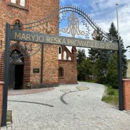 Kuta brama z napisem 'Maryjo Reska Prowadź Lud Boży' prowadząca do ceglanego kościoła z brukowanym placem i fragmentem metalowego ogrodzenia