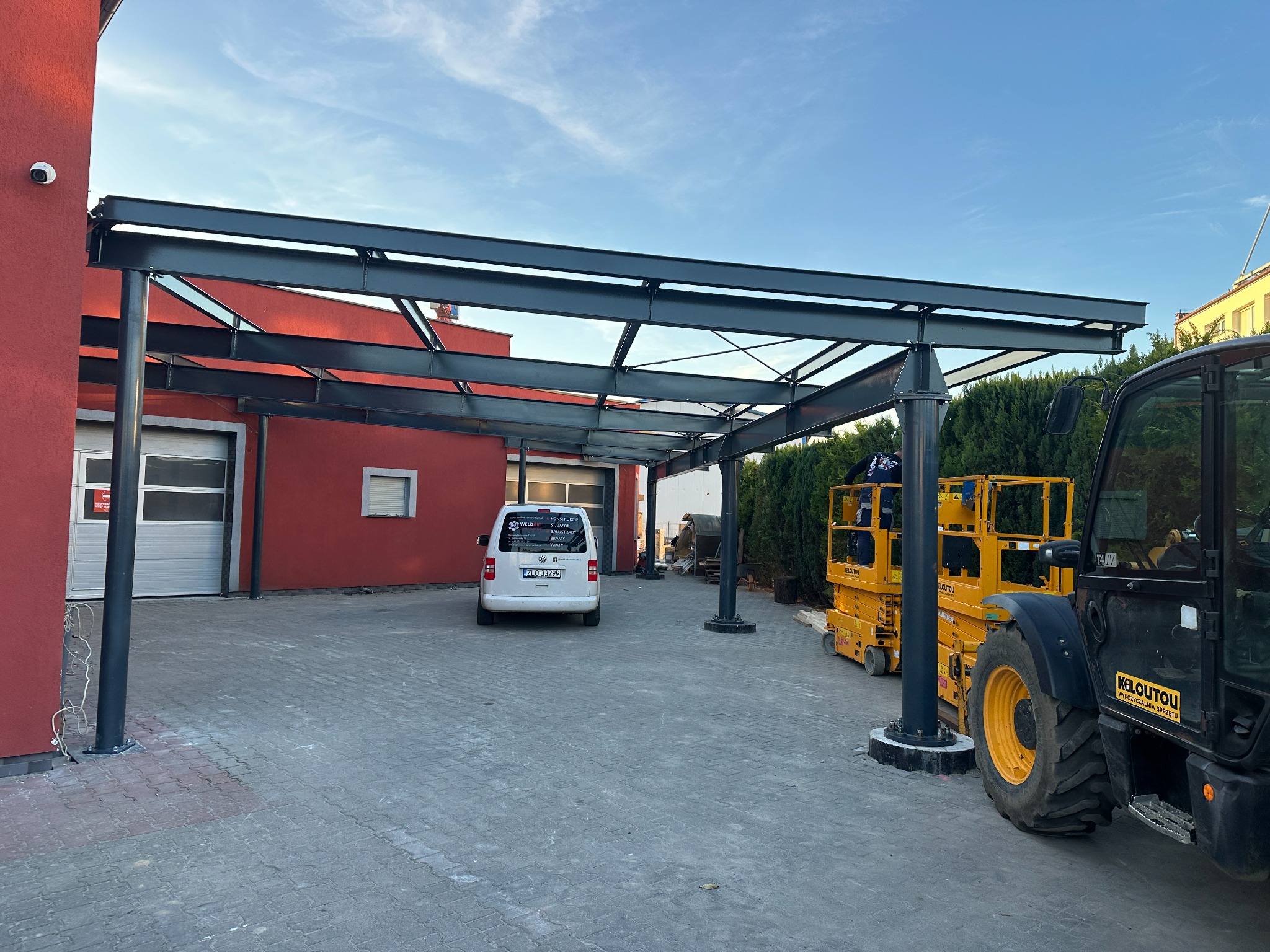Zadaszenie ze stalową konstrukcją na posesji z budynkiem w kolorze czerwieni, widoczny podnośnik koszowy i samochód dostawczy z reklamą firmy.