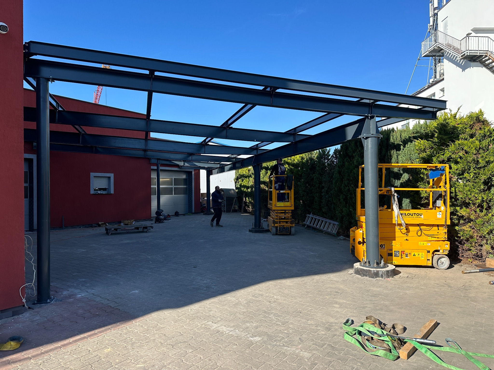 Konstrukcja stalowa z belek i słupów, prawdopodobnie zadaszenie, w trakcie montażu przy użyciu podnośników nożycowych, na placu budowy z widocznym budynkiem i zielenią w tle.
