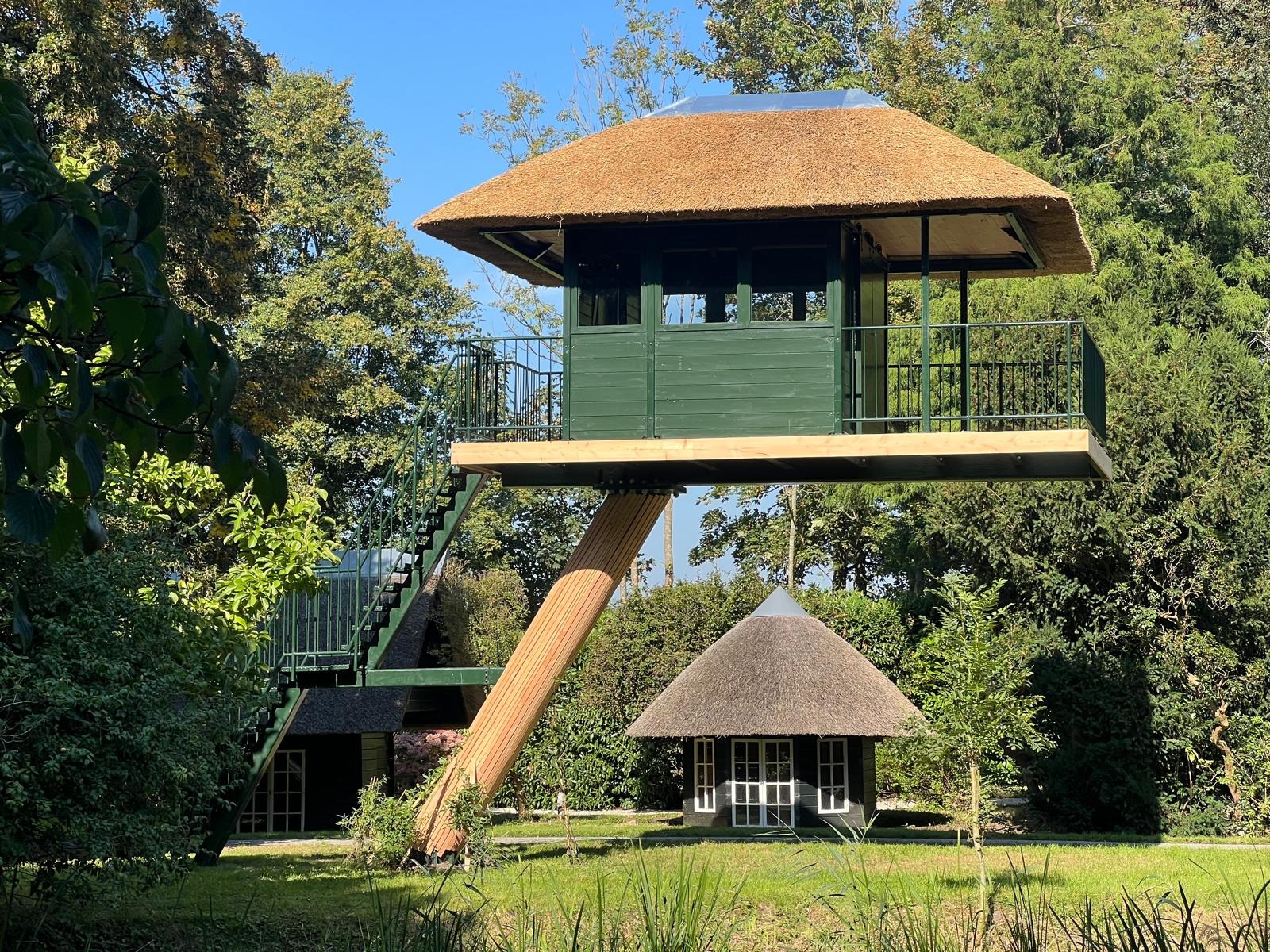 Nietypowa konstrukcja: zielona wieża widokowa z balustradą i strzechą, wsparta na drewnianym słupie, obok mniejszy budynek ze strzechą, otoczone zielenią.