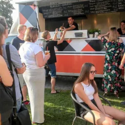 Kolejka klientów przed food truckiem z burgerami i panini, mężczyzna podnosi burgera do zdjęcia, w tle menu wypisane kredą.