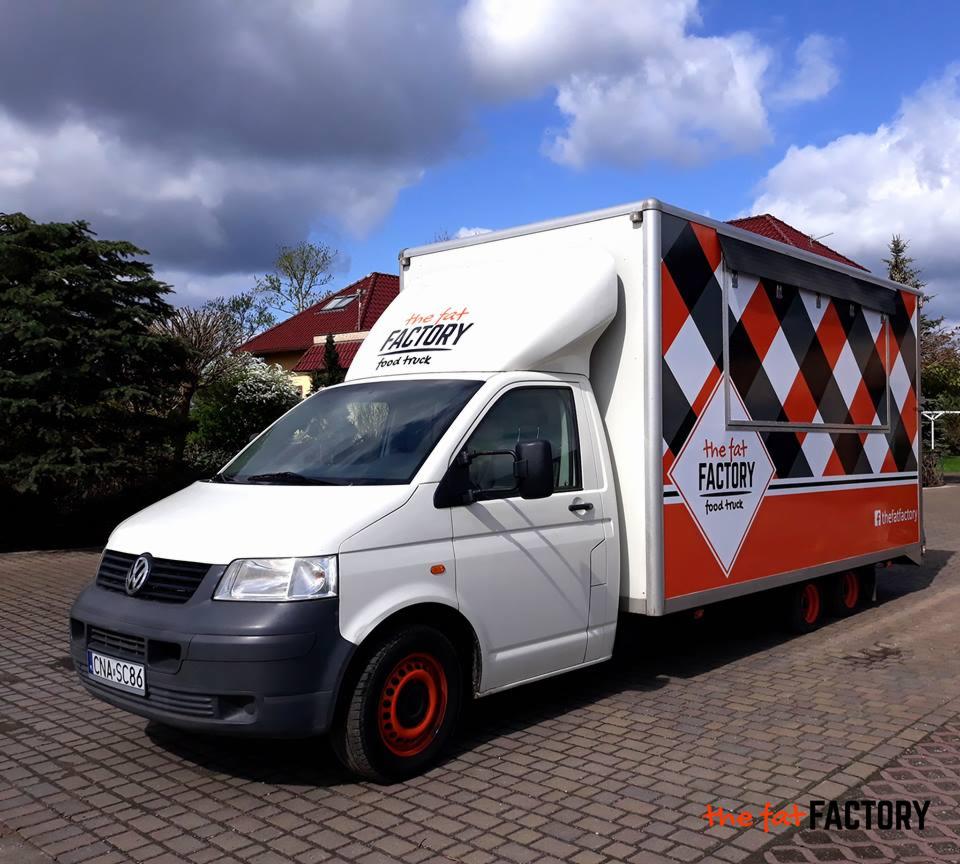 Biała furgonetka z nadrukowanym logo 'The Fat Factory food truck' na tle kostki brukowej i zieleni, z pomarańczowymi felgami i wzorem w romby na zabudowie.