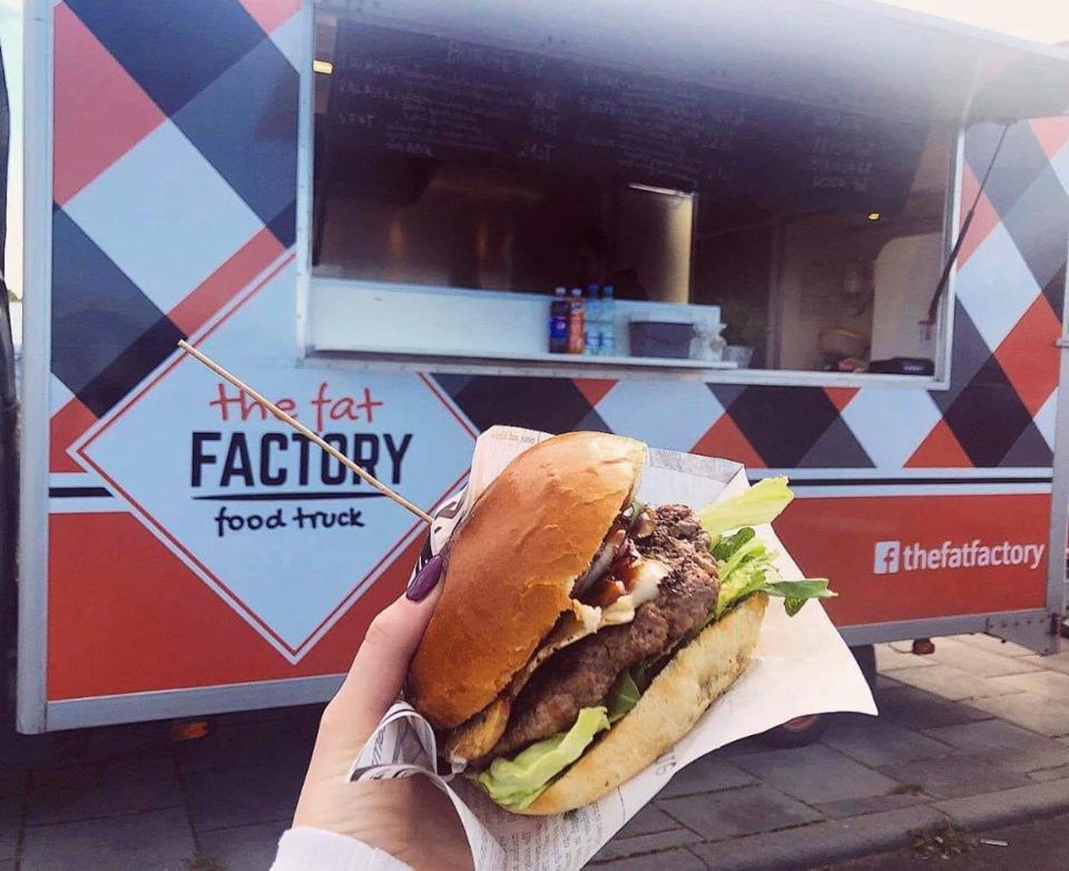 Burger trzymany w dłoni na tle food trucka z logo 'The Fat Factory', widoczny fragment menu i butelki z napojami w okienku.