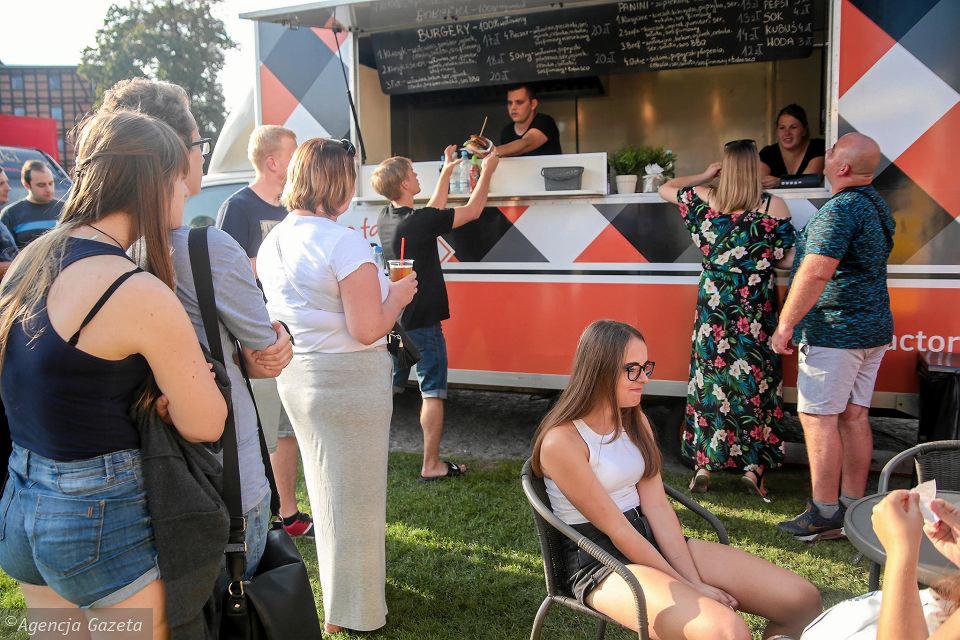 Kolejka klientów przed food truckiem z burgerami i panini, mężczyzna podnosi burgera do zdjęcia, w tle menu wypisane kredą.