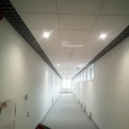 Długi, pusty korytarz z białymi ścianami i sufitem kasetonowym z oświetleniem punktowym, widok perspektywiczny, w tle okno, na pierwszym planie fragment ulotki.