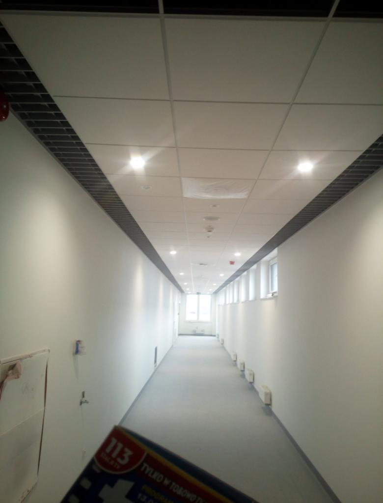 Długi, pusty korytarz z białymi ścianami i sufitem kasetonowym z oświetleniem punktowym, widok perspektywiczny, w tle okno, na pierwszym planie fragment ulotki.