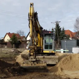 Żółta koparka w trakcie prac ziemnych, widoczny wykop w piaszczystej glebie, w tle zabudowa jednorodzinna i słupy energetyczne.