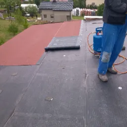 Prace dekarskie na dachu: widoczny fragment dachu pokryty papą termozgrzewalną, częściowo w kolorze czerwonym, obok butla gazowa i fragment roboczego ubrania dekarza z naszywką na kolanie.