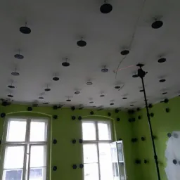 Przygotowanie do montażu płyty karton gips: zielone ściany z rozmieszczonymi uchwytami, sufit z zamocowanymi elementami mocującymi, widoczny laser poziomujący.