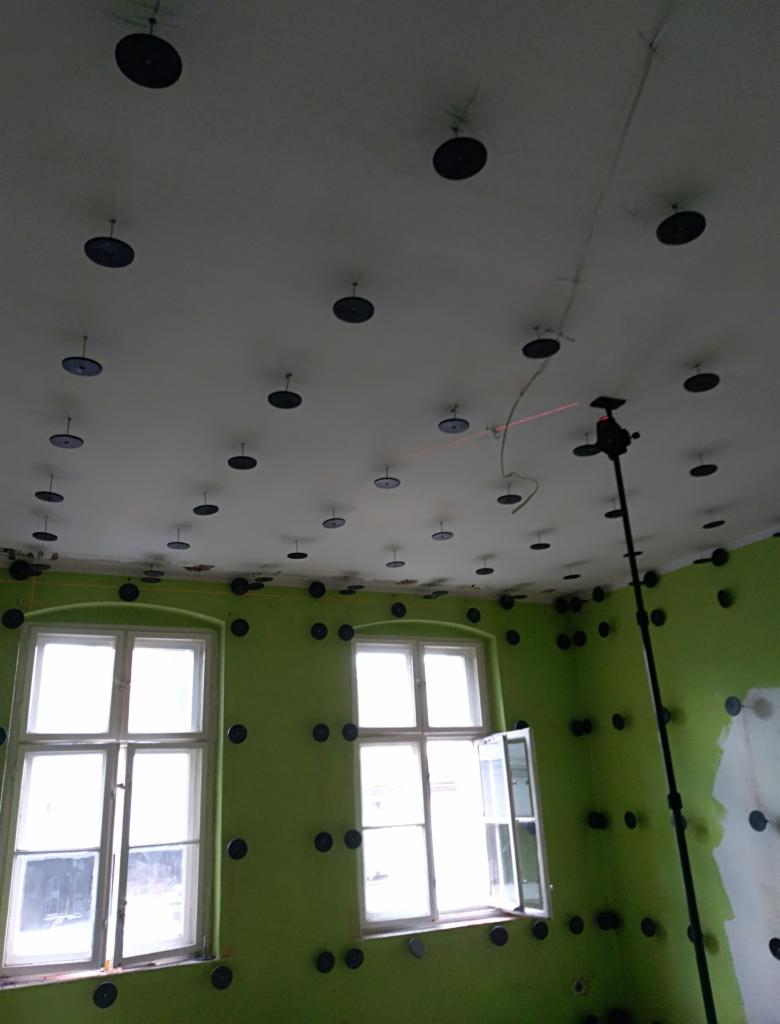 Przygotowanie do montażu płyty karton gips: zielone ściany z rozmieszczonymi uchwytami, sufit z zamocowanymi elementami mocującymi, widoczny laser poziomujący.