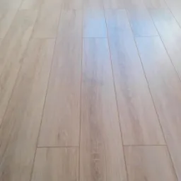 Jasne panele podłogowe ułożone wzdłuż ściany, widoczne delikatne usłojenie drewna i listwa przypodłogowa w kolorze ściany.