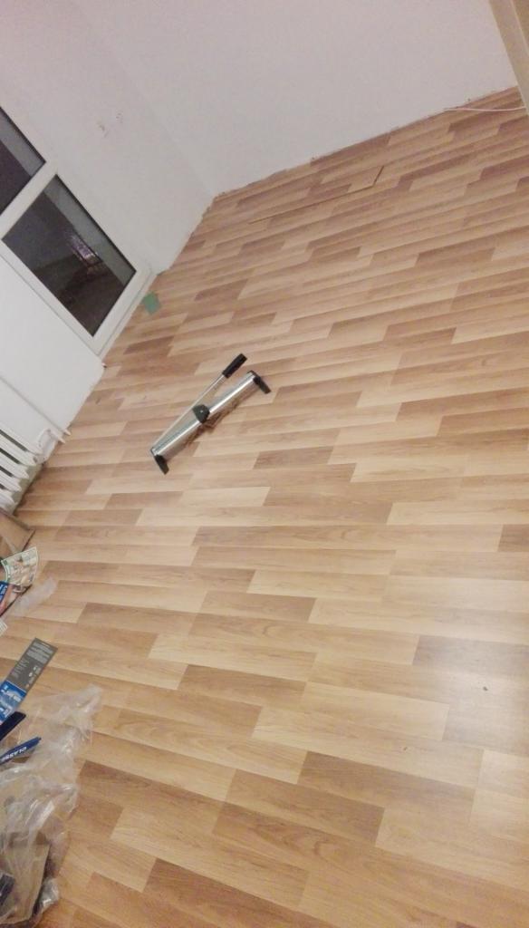 Fragment pokoju z ułożonymi panelami podłogowymi o wzorze drewna i narzędziem do dociskania paneli, widoczne niedociągnięcia przy ścianie.