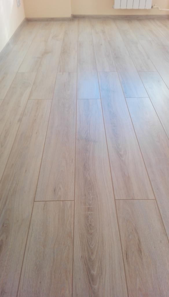 Jasne panele podłogowe ułożone wzdłuż ściany, widoczne delikatne usłojenie drewna i listwa przypodłogowa w kolorze ściany.