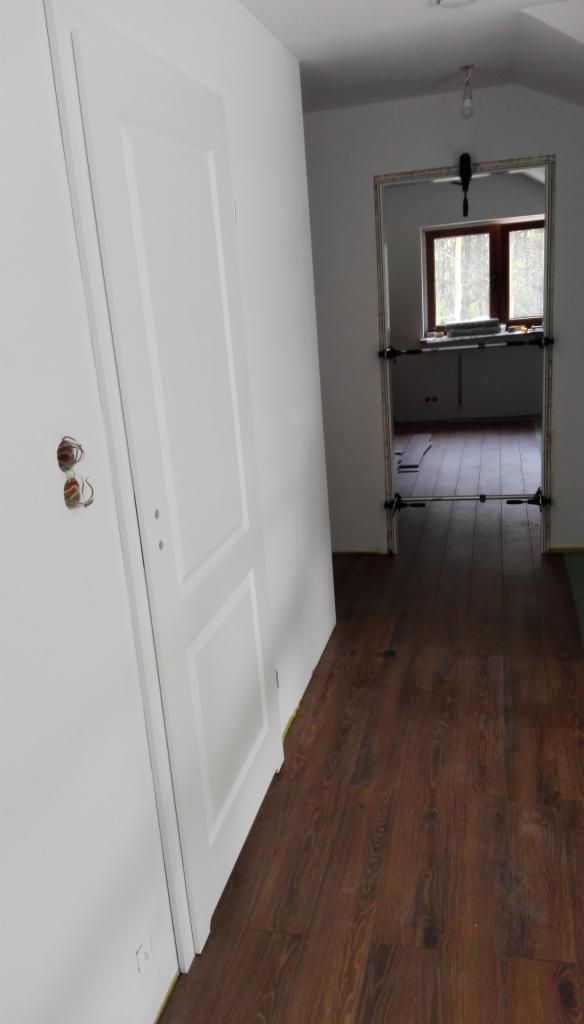 Białe, częściowo zamontowane drzwi wewnętrzne z widocznymi przewodami elektrycznymi w ścianie, brązowe panele podłogowe i widok na okno w tle przez otwór drzwiowy wzmocniony metalowymi rozpórkami.