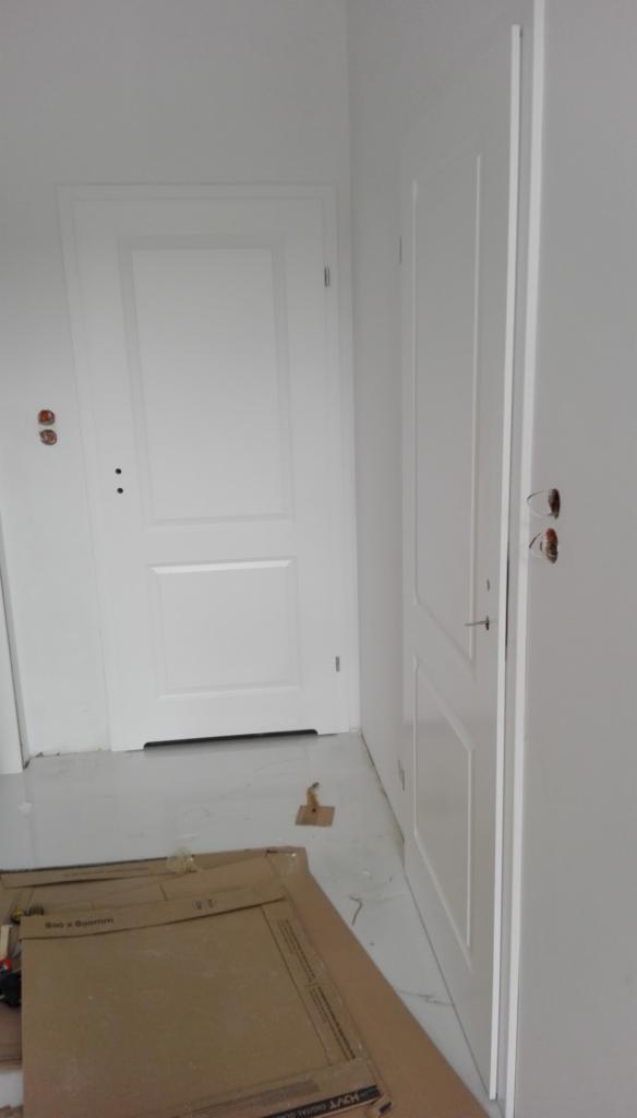 Dwie białe, klasyczne drzwi wewnętrzne, jedna zamontowana, druga oparta o ścianę w trakcie montażu, widoczne kartony po panelach na podłodze.