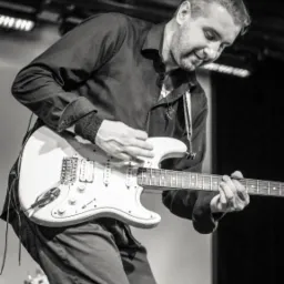 Czarno-białe zdjęcie muzyka grającego na białej gitarze elektrycznej, z perkusistą w tle i oświetleniem scenicznym nad głową.