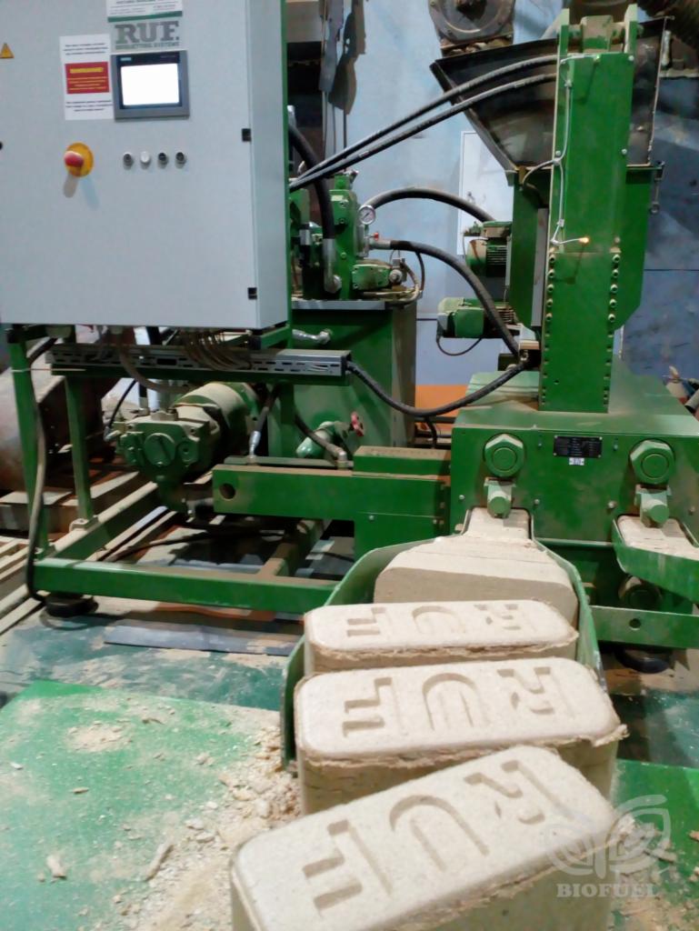 Proces produkcji brykietu: zielona maszyna z szarym panelem sterowania RUF formuje gotowe kostki z napisem RUF, widoczne trociny na podłodze.