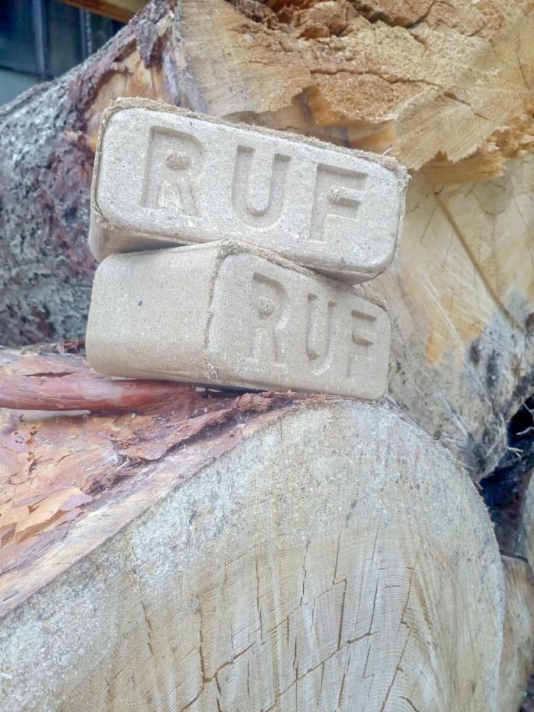 Dwa brykiety drzewne RUF ułożone jeden na drugim na tle stosu drewna opałowego z widocznymi słojami.