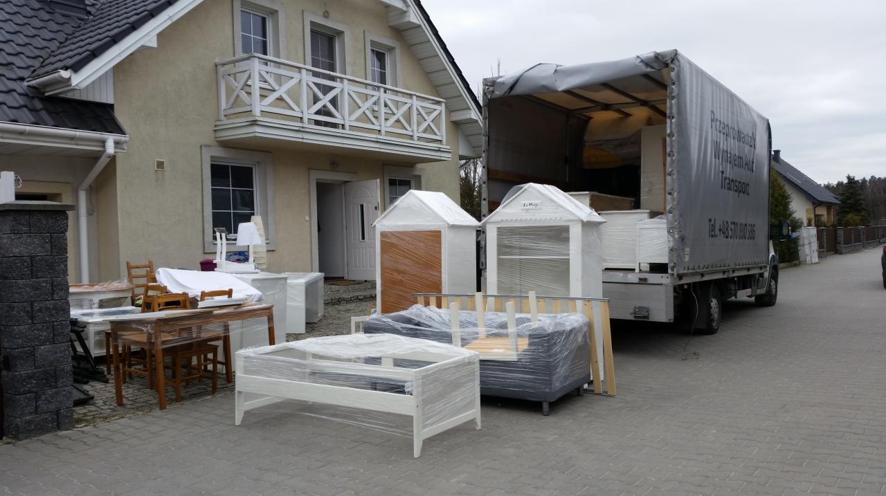 Meble zabezpieczone folią, częściowo załadowane do ciężarówki transportowej przed domem z balkonem.