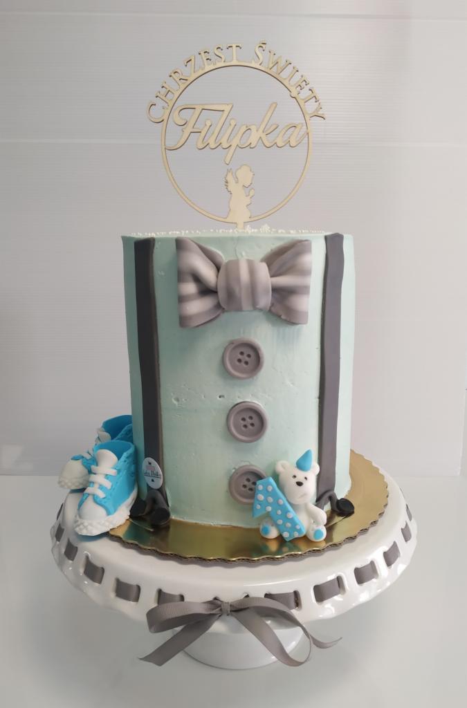 Tort na Chrzest Święty Filipka, zdobiony motywem ubranka z muchą, guzikami, szelkami, małymi bucikami i misiem z cyfrą 1 na torcie, całość na białym stojaku.