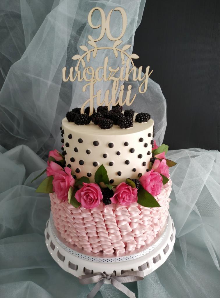 Dwupiętrowy tort urodzinowy z różową falbanką, białym lukrem w czarne kropki, dekorowany jeżynami i różami, z drewnianym topperem '90 urodziny Julii'.