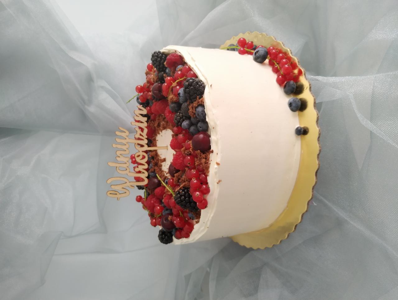Tort urodzinowy z białym kremem, dekorowany świeżymi czerwonymi porzeczkami, jeżynami i borówkami, z drewnianym napisem 'W dniu urodzin', na złotym podkładzie i tle z delikatnej tkaniny.