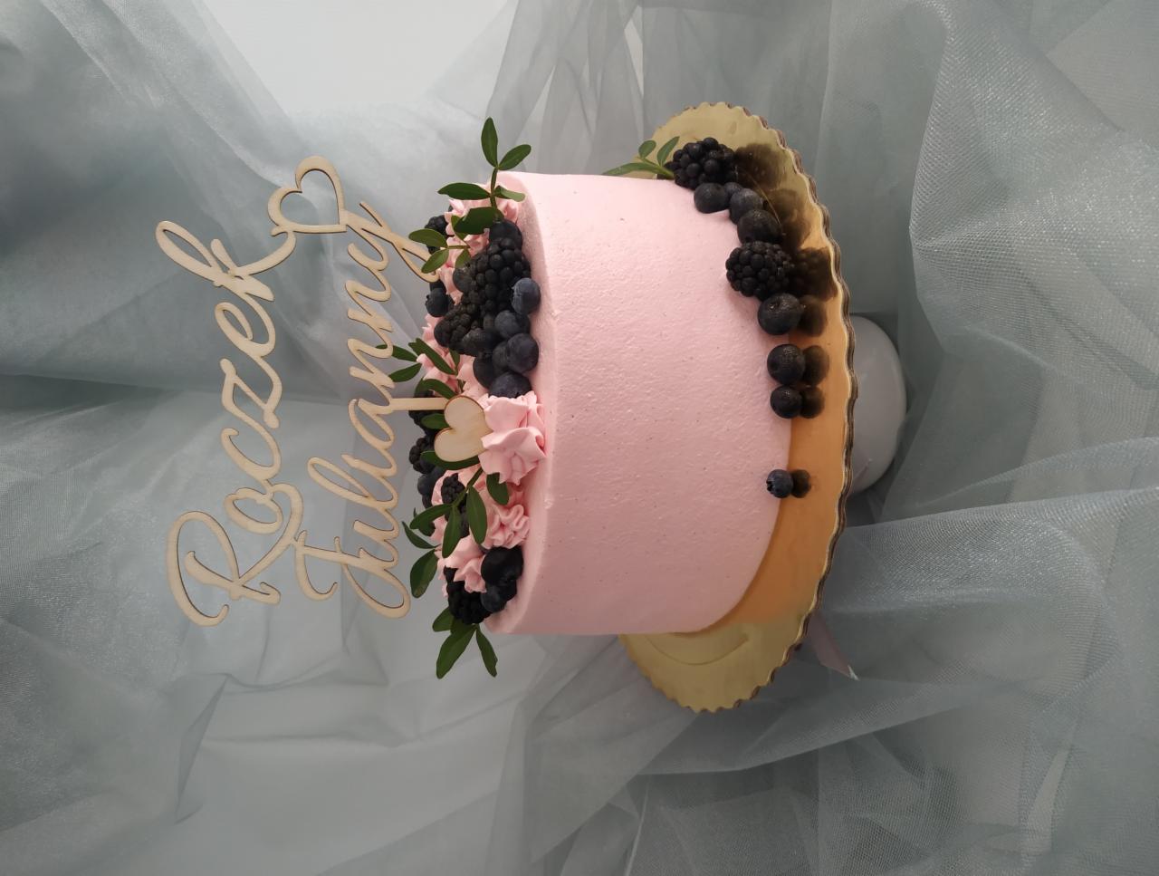 Tort urodzinowy dla Julianny z różowym kremem, borówkami, jeżynami i drewnianym napisem 'Roczek Julianny' na złotym podkładzie, tło z szarego tiulu.