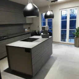 Nowoczesna kuchnia z wyspą i minimalistycznymi lampami wiszącymi, widok na okno z rośliną doniczkową. Na wyspie ułożone szare pudełka.