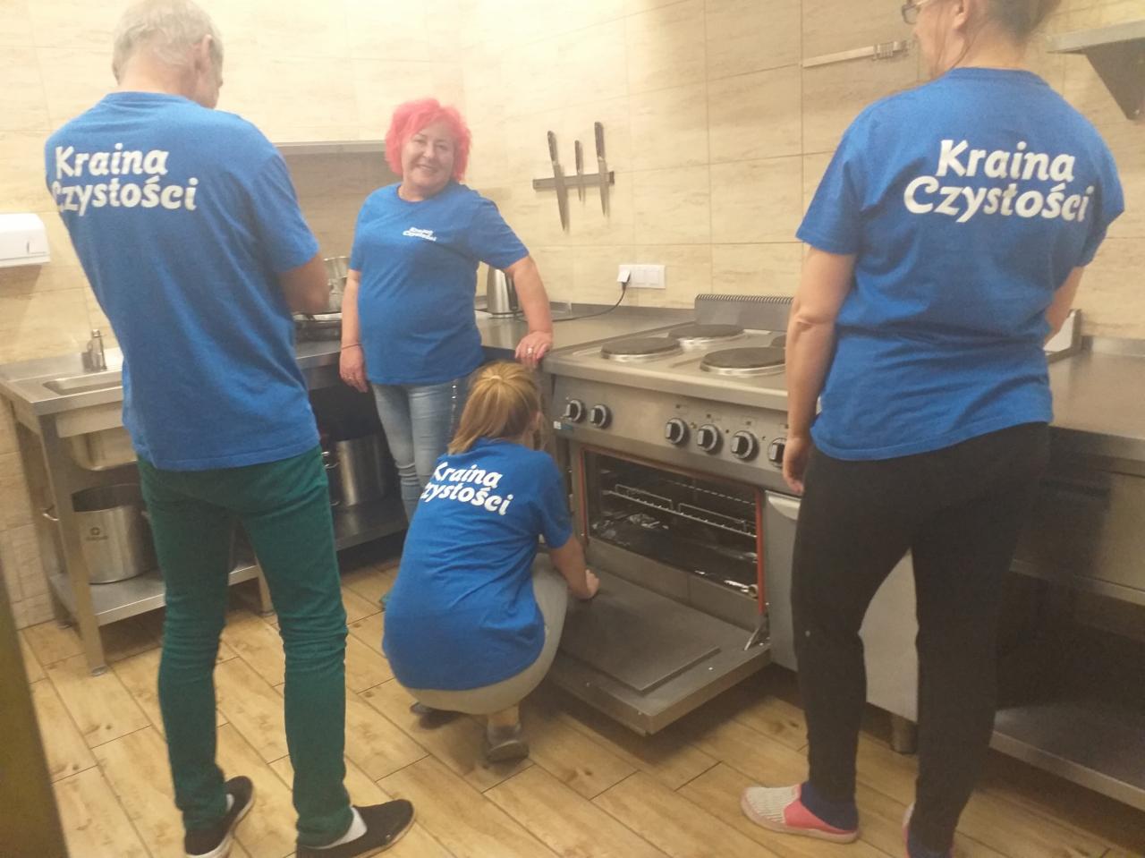 Zespół sprzątający w niebieskich koszulkach z napisem 'Kraina Czystości' czyści wnętrze piekarnika w kuchni z płytkami. Na ścianie wiszą dekoracyjne noże.