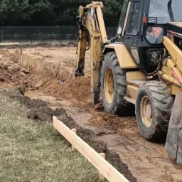 Żółta koparko-ładowarka CAT wykopuje wąski wykop pod fundamenty, widoczny pryzma ziemi i drewniane szalunki.