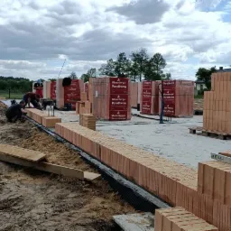 Wznoszenie muru z czerwonej ceramiki Porotherm na fundamencie z izolacją pionową; murarze przy pracy, w tle palety z cegłami i zielone drzewa.