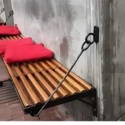 Metalowa ławka podwieszana do ściany z drewnianym siedziskiem i czerwonymi poduszkami, przymocowana za pomocą kutego metalowego wspornika.