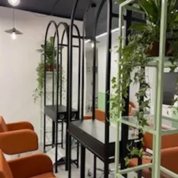 Nowoczesne stanowiska fryzjerskie z metalowymi konstrukcjami w kształcie łuków, wyposażone w lustra i półki, ozdobione roślinami doniczkowymi, z pomarańczowymi fotelami w tle.