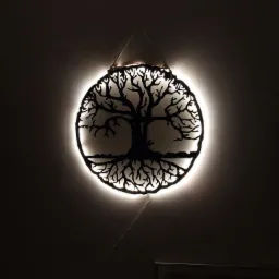 Dekoracja ścienna: metalowy ażurowy okrąg z motywem drzewa podświetlony od tyłu ciepłym białym światłem, zawieszona na sznurku na ciemnoszarej ścianie.