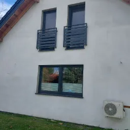 Elewacja domu z dwoma małymi balkonami z czarnymi, poziomymi barierkami, okno z żaluzjami i klimatyzator na trawniku.