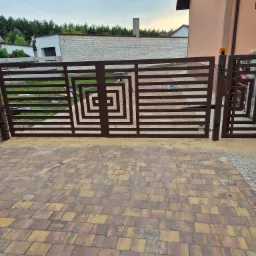 Nowoczesna brama wjazdowa w kolorze brązowym, z geometrycznym wzorem kwadratów. Widoczny automat do bramy i kostka brukowa w odcieniach brązu i żółci.