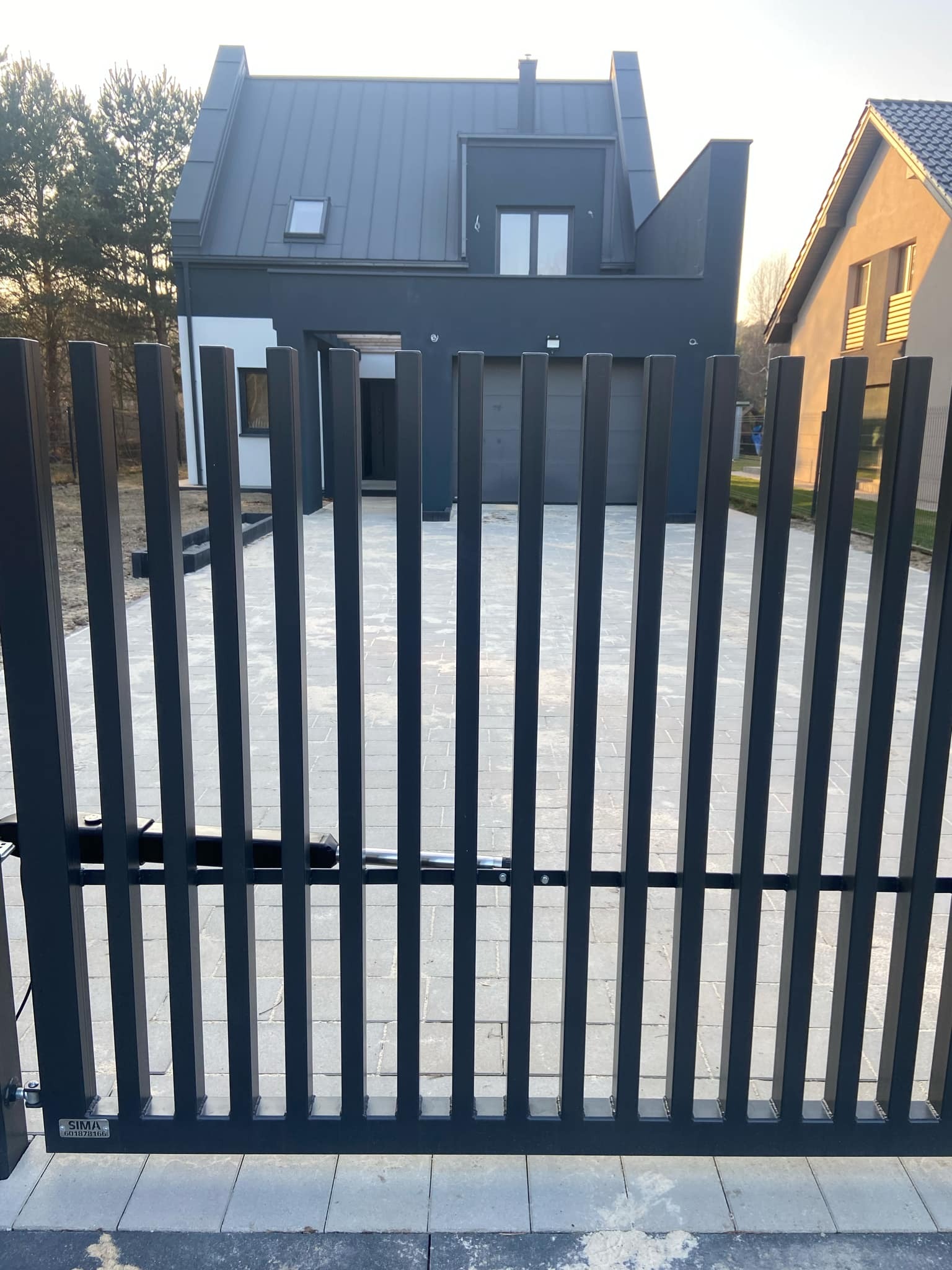 Nowoczesna, automatyczna brama wjazdowa z pionowymi szczeblami, widoczny mechanizm otwierający, w tle dom w minimalistycznym stylu z szarą elewacją i brukowany podjazd.