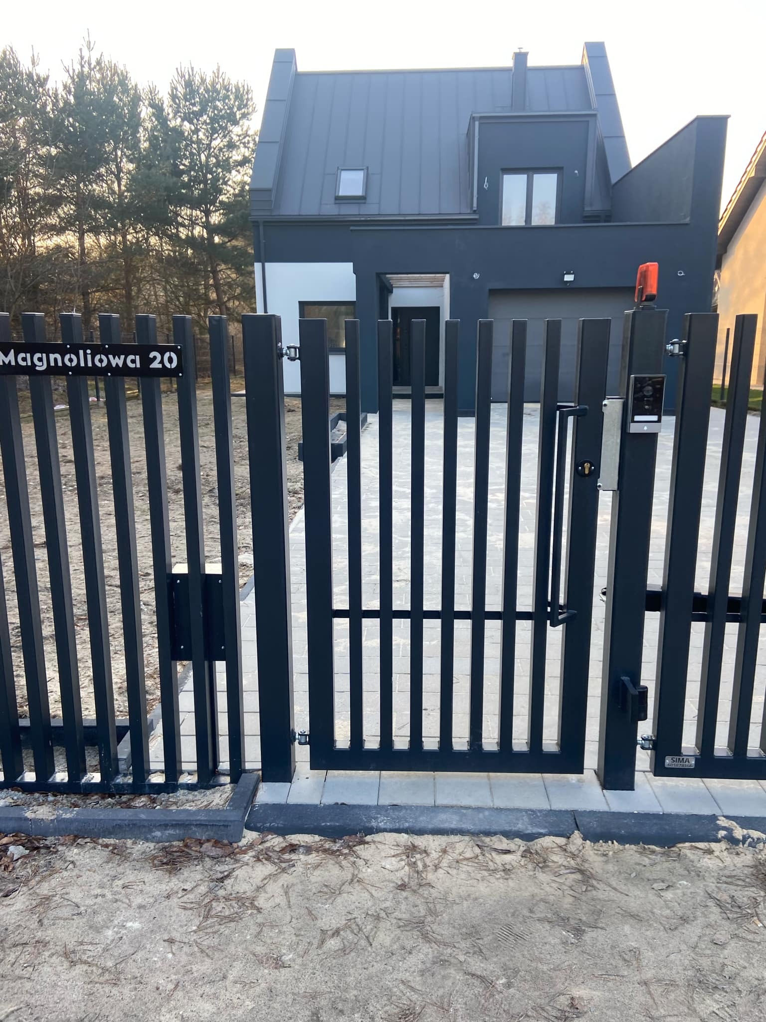 Nowoczesna, grafitowa brama wjazdowa z pionowymi sztachetami i domofonem przy domu z szarą elewacją i dachem, tabliczka z adresem 'Magnoliowa 20'.