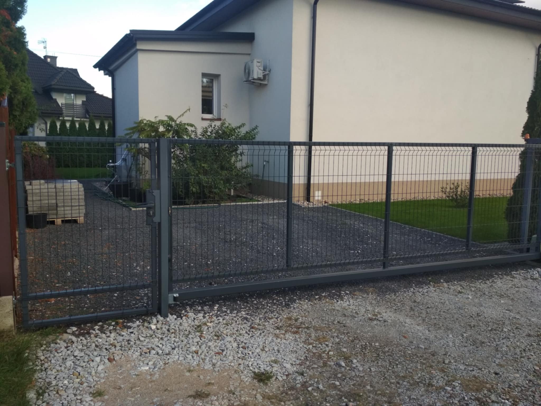 Przesuwna brama wjazdowa z metalowej siatki na posesji, widoczny dom z klimatyzacją i paleta kostki brukowej w tle.