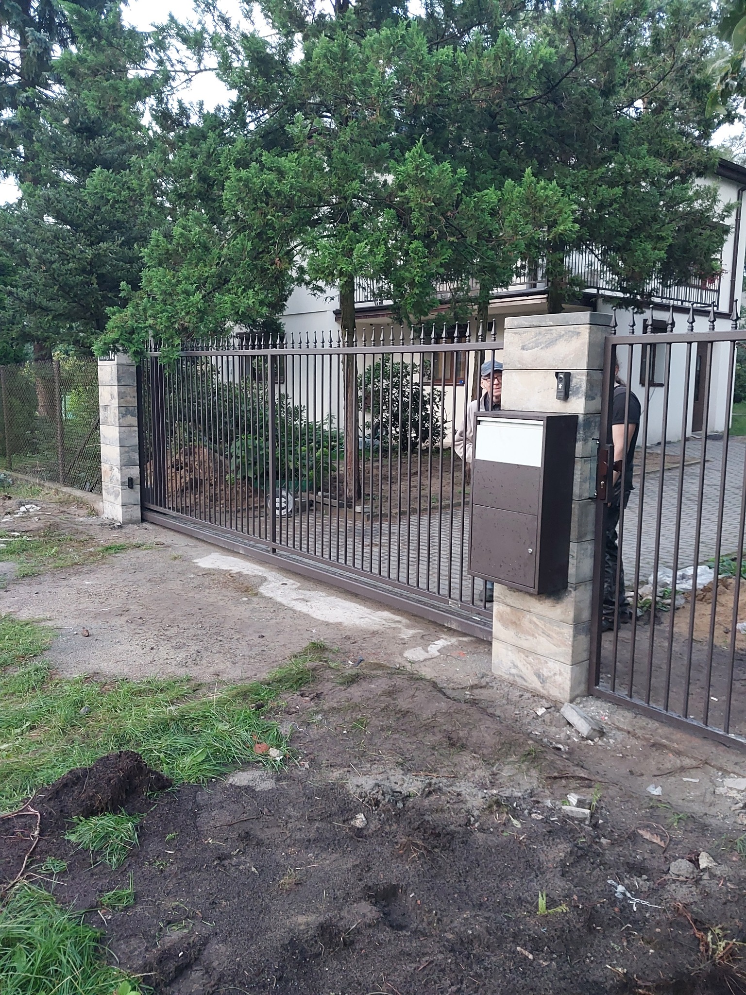 Przesuwna brama wjazdowa z metalowymi prętami, zamontowana na kamiennych słupkach z domofonem i skrzynką na listy, widoczna na tle domu i zieleni, teren wokół w trakcie prac wykończeniowych.