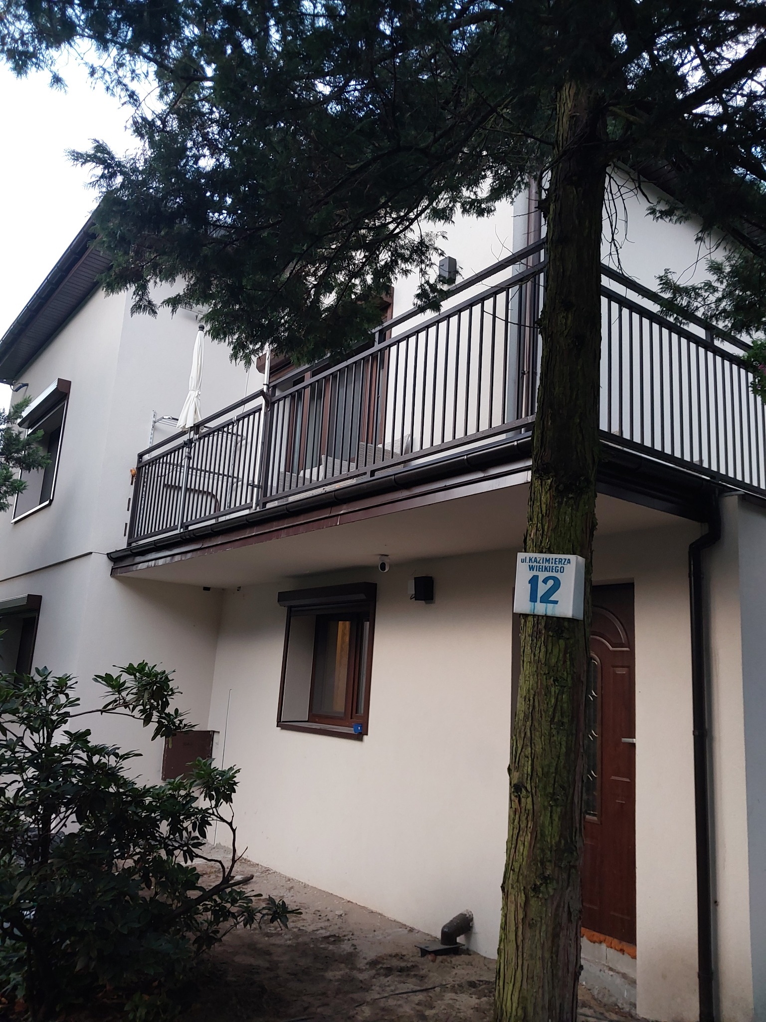 Balkon z czarną metalową balustradą, widok z dołu, na drzewie tabliczka z adresem.
