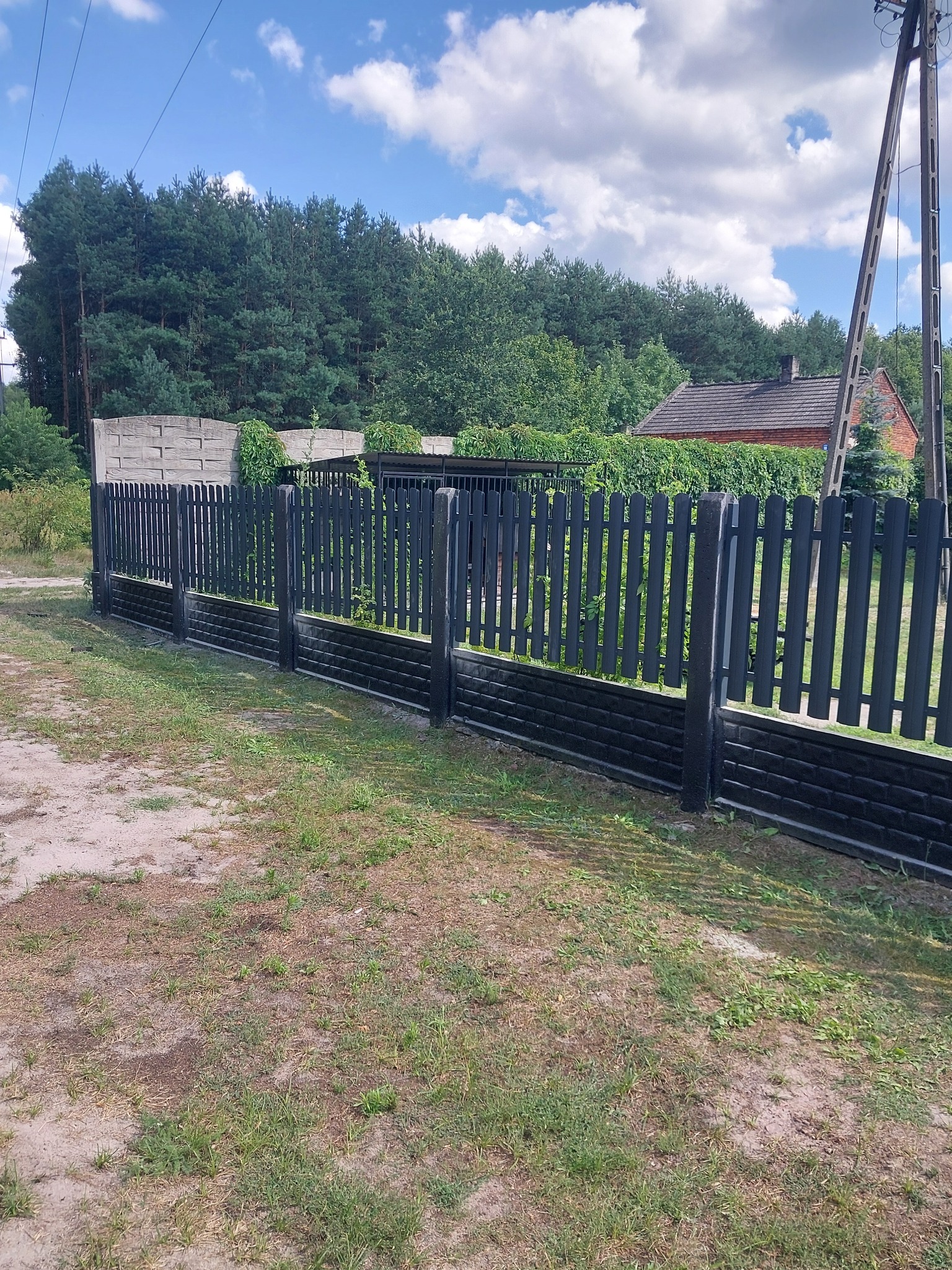 Ciemne ogrodzenie panelowe z elementami imitującymi cegłę, oddzielające posesję od drogi gruntowej z widokiem na dom i las w tle.