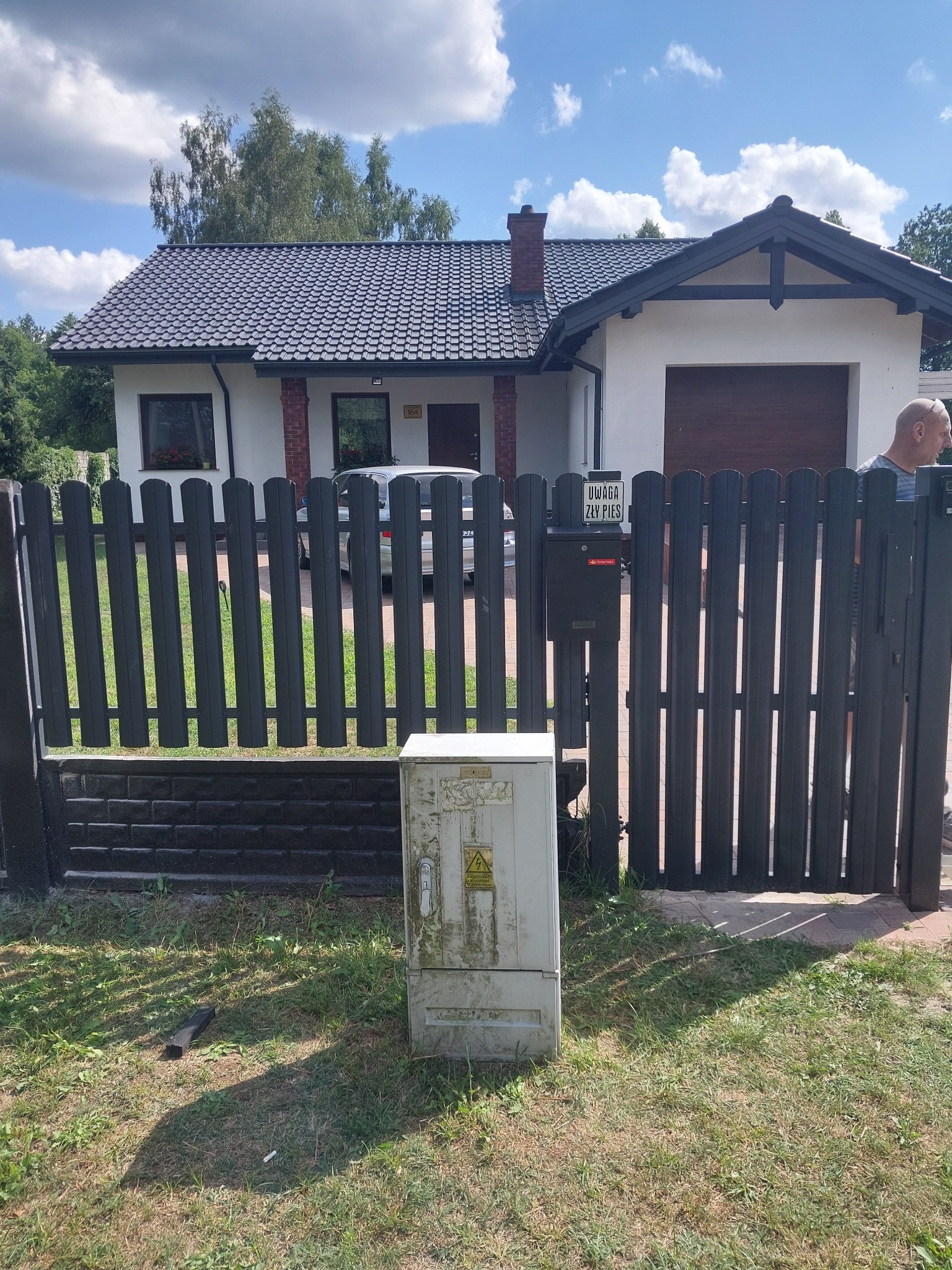 Ciemnoszare ogrodzenie panelowe z metalowymi sztachetami i elementami imitującymi cegłę, przed domem z garażem i tabliczką 'Uwaga zły pies'.