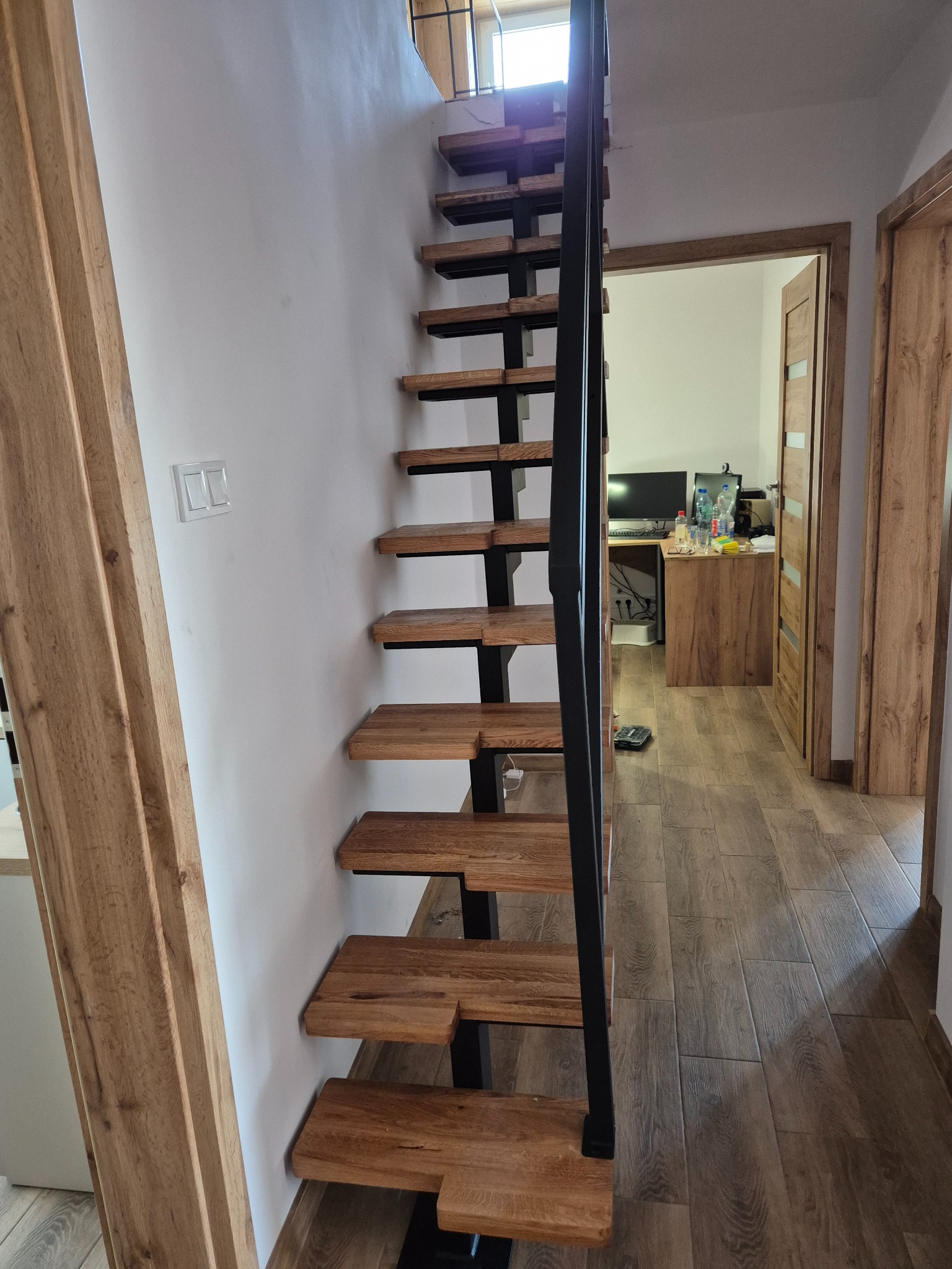 Wąskie, drewniane schody metalowe bez balustrady, zamontowane przy ścianie w jasnym wnętrzu, widoczne z dołu, z drewnianymi panelami podłogowymi i drzwiami w tle.