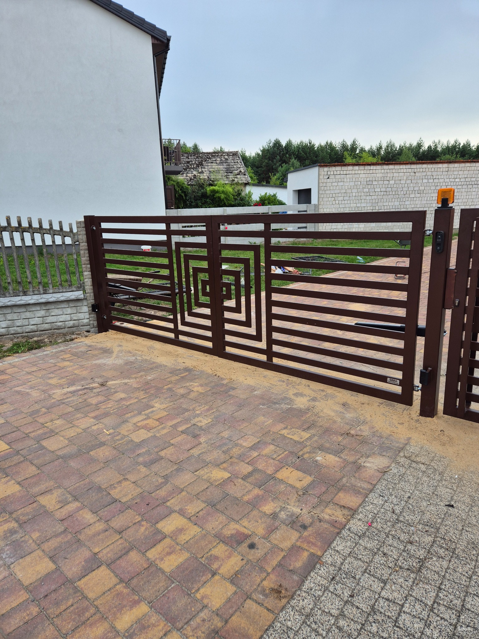Nowoczesna brama wjazdowa z poziomych profili w kolorze brązowym, z geometrycznym wzorem w centralnej części. Kostka brukowa przed bramą. Widok na dom i zieleń.