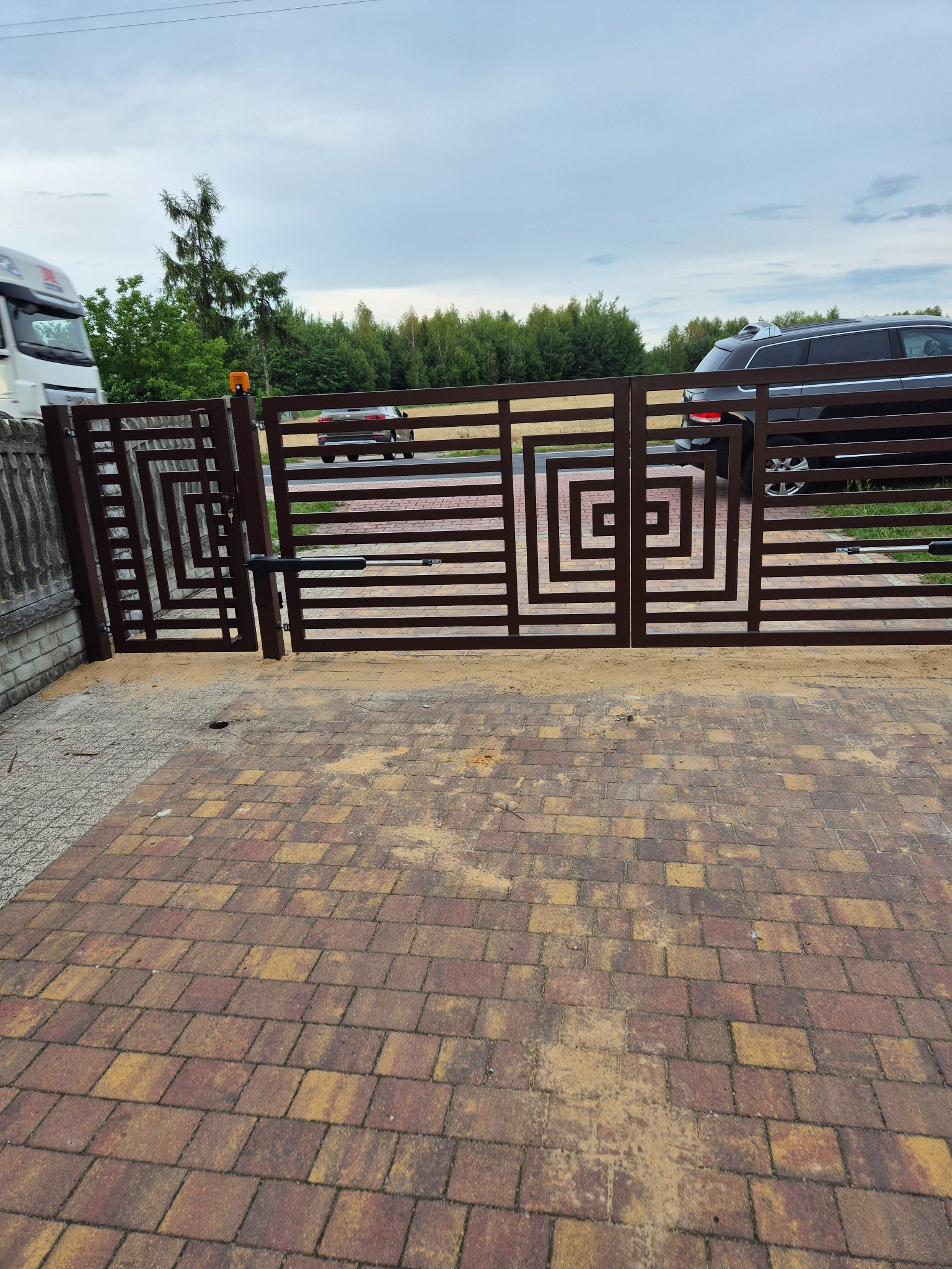 Nowoczesna, brązowa brama wjazdowa z automatyką, o geometrycznym wzorze, na tle kostki brukowej i zieleni. Widoczny samochód osobowy i ciężarówka.