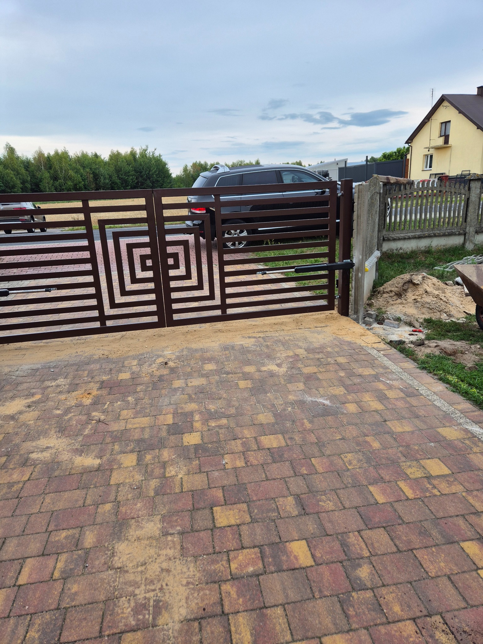 Nowoczesna, brązowa brama wjazdowa z automatyką, z geometrycznym wzorem na tle domu i samochodu. Brama częściowo otwarta, widoczne elementy montażu.